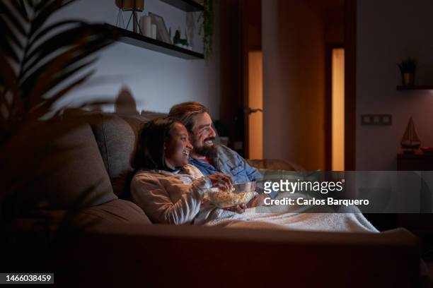 mixed race young couple at home watching movies at night in the sofa and eating popcorns. - movie and tv fotos stock-fotos und bilder