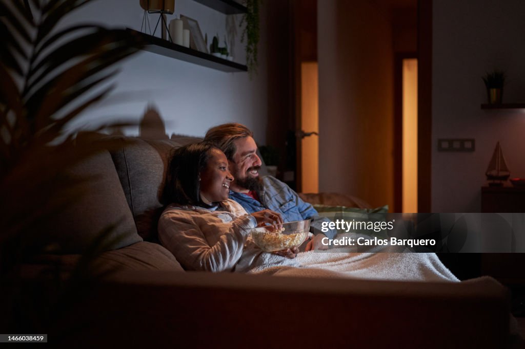 Mixed race young couple at home watching movies at night in the sofa and eating popcorns.
