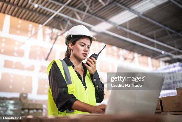 junge frau, die auf dem walkie-talkie spricht, während sie den laptop in einem lagerhaus benutzt - bauingenieurwesen stock-fotos und bilder