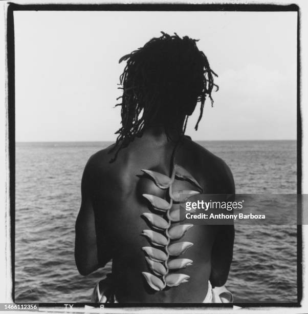 Portrait, from behind, of an unidentied model posing, topless with a heliconia plant on their back, overlookingthe water, Jamaica, 1997.
