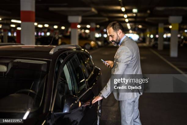 empresário de meia-idade entrando em seu carro e usando telefone na garagem pública. - entrando - fotografias e filmes do acervo