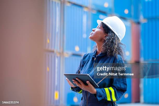 cargo container field inspection with mobile application. female african american dock worker holding a tablet while inspecting the packaging of the goods and recording loading conditions and compiling a report in a shipping yard. - macacão preto imagens e fotografias de stock