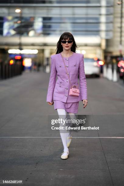 Mary Leest wears black sunglasses, diamond earrings, a pale purple shoulder-pads / buttoned jacket, a matching pale purple tweed short skirt, a pale...