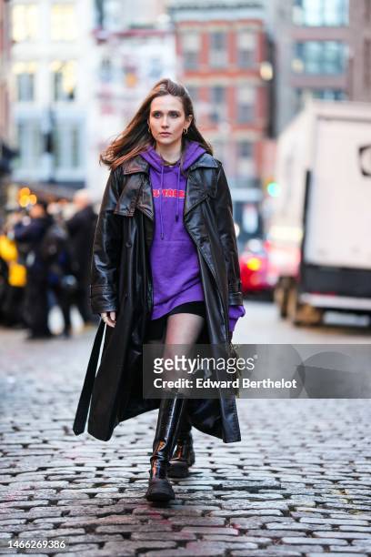 Iana Rad wears gold earrings, a dark purple with red print pattern and embroidered rhinestones hoodie sweater, a black shiny leather long coat, black...