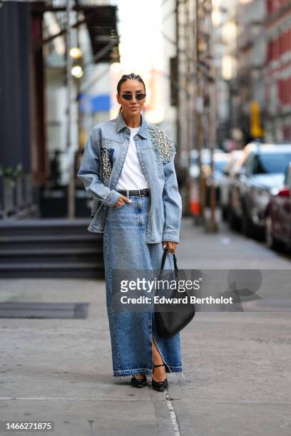 Tamara Kalinic wears black circle sunglasses, gold earrings, a white t-shirt, a black shiny leather belt, a blue faded denim with embroidered large...