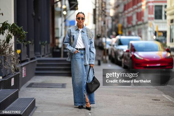 Tamara Kalinic wears black circle sunglasses, gold earrings, a white t-shirt, a black shiny leather belt, a blue faded denim with embroidered large...