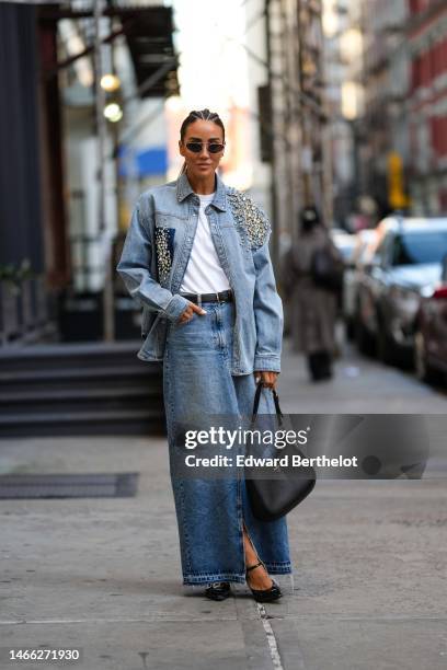 Tamara Kalinic wears black circle sunglasses, gold earrings, a white t-shirt, a black shiny leather belt, a blue faded denim with embroidered large...