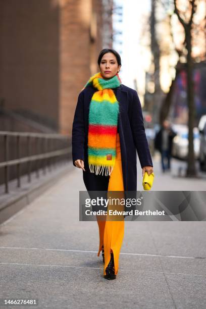 Samia Laaboudi wears gold earrings, a neon orange / yellow / red / green striped print pattern fluffy wool large scarf from Loewe, a navy blue long...