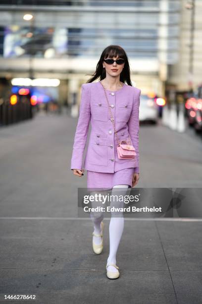 Mary Leest wears black sunglasses, diamond earrings, a pale purple shoulder-pads / buttoned jacket, a matching pale purple tweed short skirt, a pale...