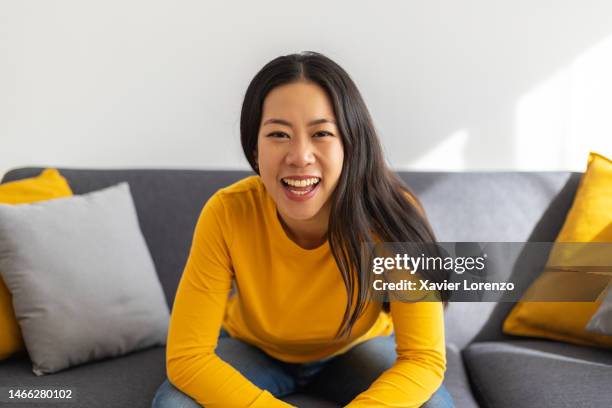 smiling portrait of cheerful asian young adult woman resting on sofa looking at camera in the living room. front view of chinese happy female relaxing at home - smiling facing camera sofa stock pictures, royalty-free photos & images