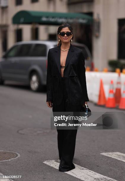 Guest wears black sunglasses, gold earrings, a gold large chain necklace, a black bra underwear, a black silk / cut-out waist blazer jacket, matching...