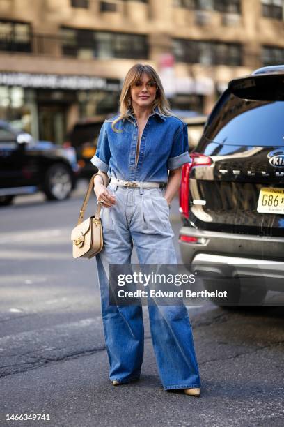 Guest wears gold glasses, gold and beige leather pendant earrings, a gold chain necklace, a navy blue denim short sleeves rolled-up shirt, a beige...