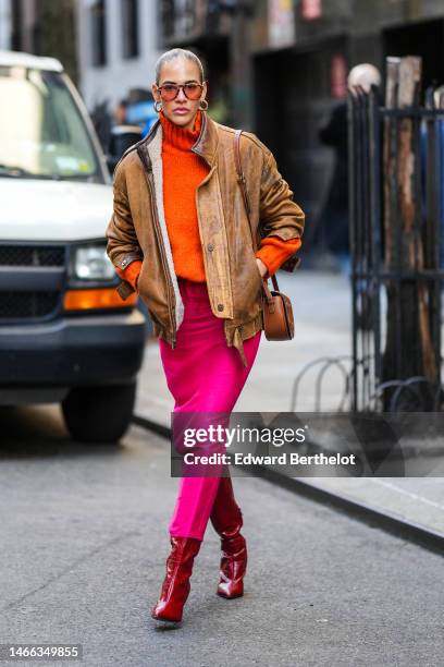 Guest wears orange sunglasses, gold earrings, a neon orange ribbed wool turtleneck pullover, a brown faded leather zipper coat, a brown shiny leather...