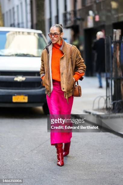 Guest wears orange sunglasses, gold earrings, a neon orange ribbed wool turtleneck pullover, a brown faded leather zipper coat, a brown shiny leather...