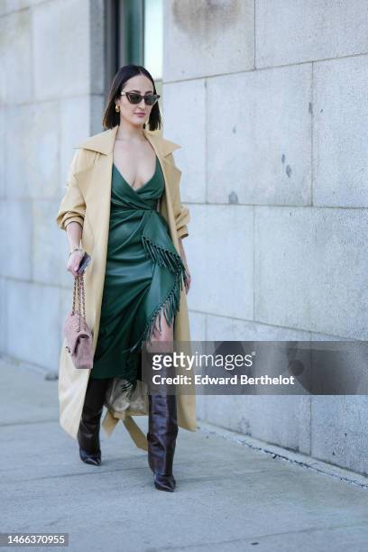 Guest wears brown sunglasses, gold earrings, a dark green shiny leather V-neck / wrap asymmetric knees dress with embroidered fringed borders, a pale...