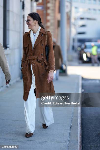 Guest wears black aviator sunglasses, gold earrings, a white long scarf, a white t-shirt, white denim wide legs pants, a dark brown suede belted long...