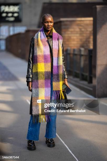 Model wears a black shiny leather jacket, a yellow and purple checkered print pattern fluffy long fringed scarf from Acne Studio, blue denim ripped...