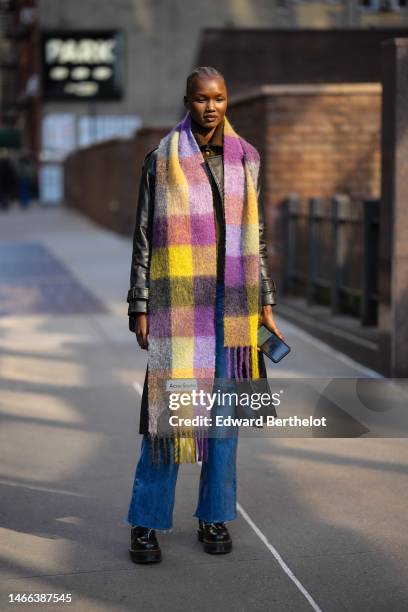 Model wears a black shiny leather jacket, a yellow and purple checkered print pattern fluffy long fringed scarf from Acne Studio, blue denim ripped...
