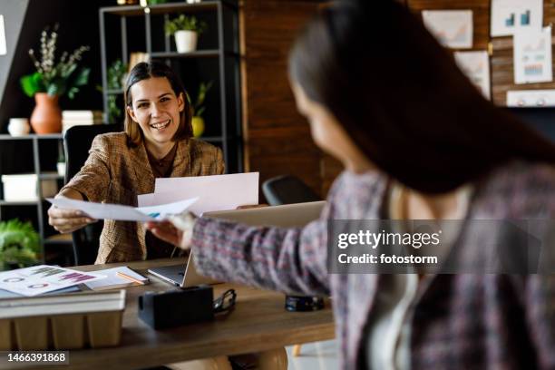 young businesswoman hanging documents to her colleague sitting at desk behind her - doorgeven stockfoto's en -beelden