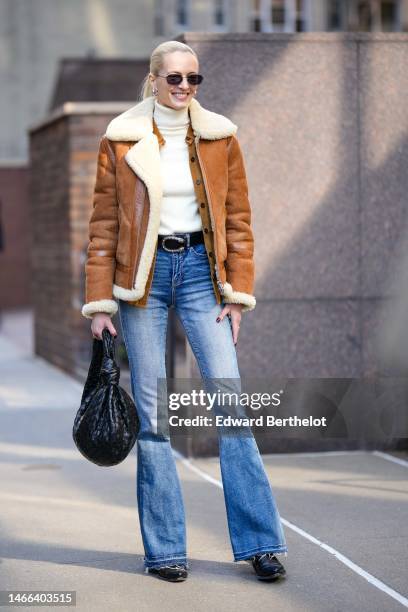 Model wears black sunglasses, a white turtleneck wool pullover, a camel suede with beige white sheep interior zipper aviator coat, a black shiny...