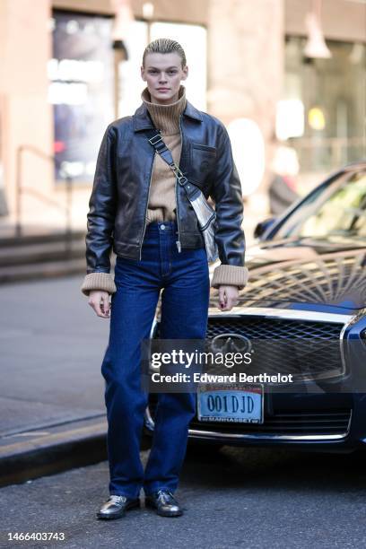 Model wears a beige ribbed wool turtleneck pullover, a black shiny leather zipper jacket, a silver shiny leather zipper crossbody bag, navy blue...