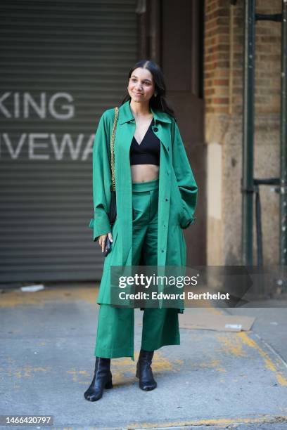 Guest wears a black V-neck cropped top, a green long coat, green suit pants, a black embossed leather shoulder bag, black shiny leather block heels /...