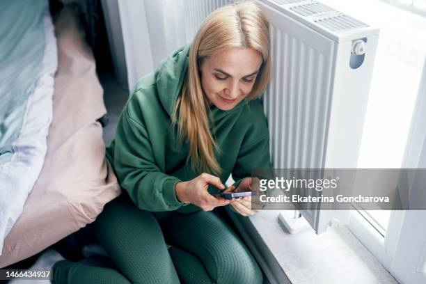 a young woman at a heating radiator with a mobile phone at home in winter . - district heating stock pictures, royalty-free photos & images