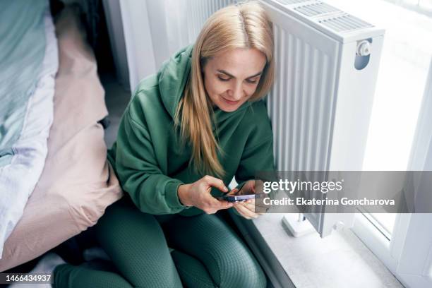 a young woman at a heating radiator with a mobile phone at home in winter . - district heating plant 個照片及圖片檔