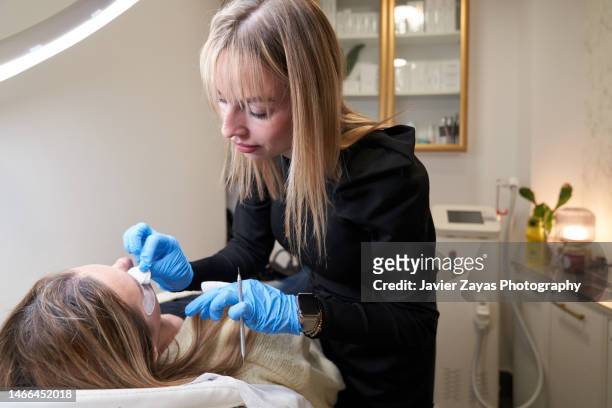 beautician doing eyelash extension process to young woman in beauty salon - eyelashes stock pictures, royalty-free photos & images