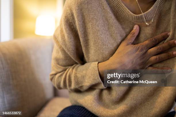 black woman experiences chest pain sitting on sofa - cardiopatía fotografías e imágenes de stock