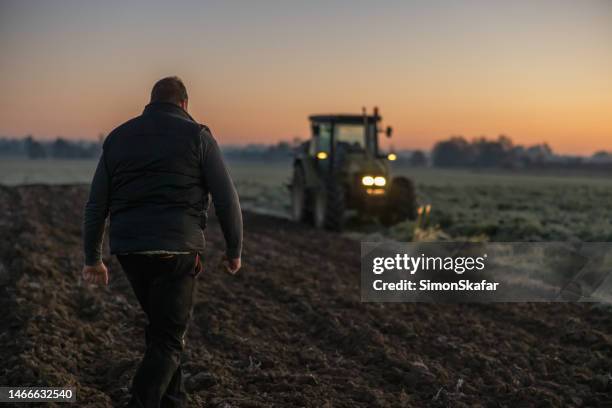 man with short brown hair,and a black vest is walking on his agricultural field in the evening,with tractor in the background,lights on tractor are turned on - tractors stock pictures, royalty-free photos & images