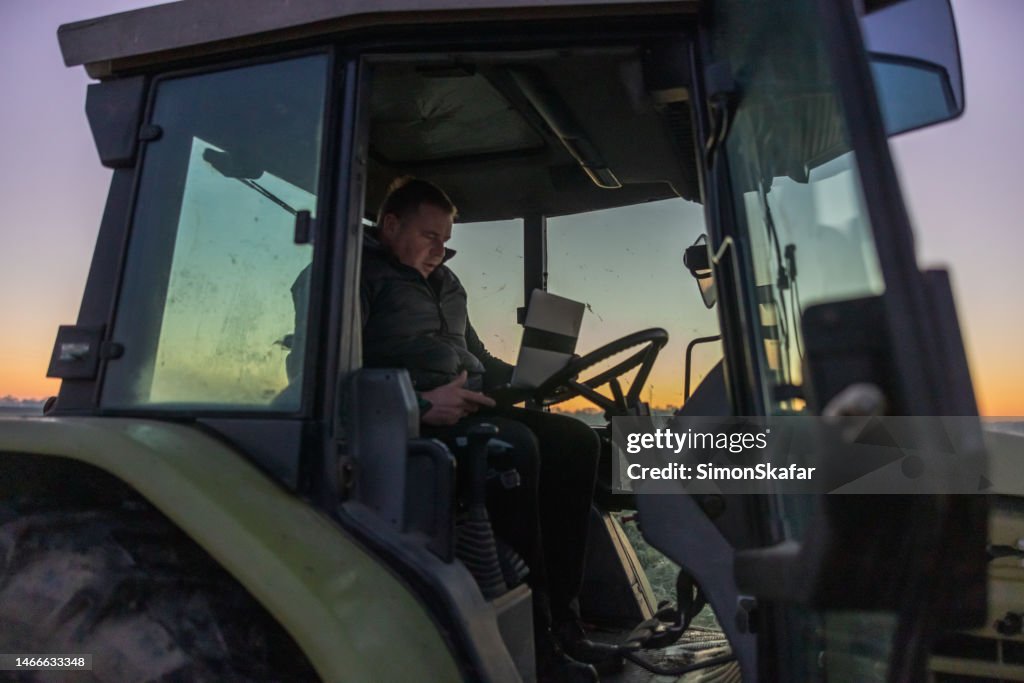 Man with short brown hair,black vest and trousers sitting inside his tractor with a laptop in the evening,working on the laptop