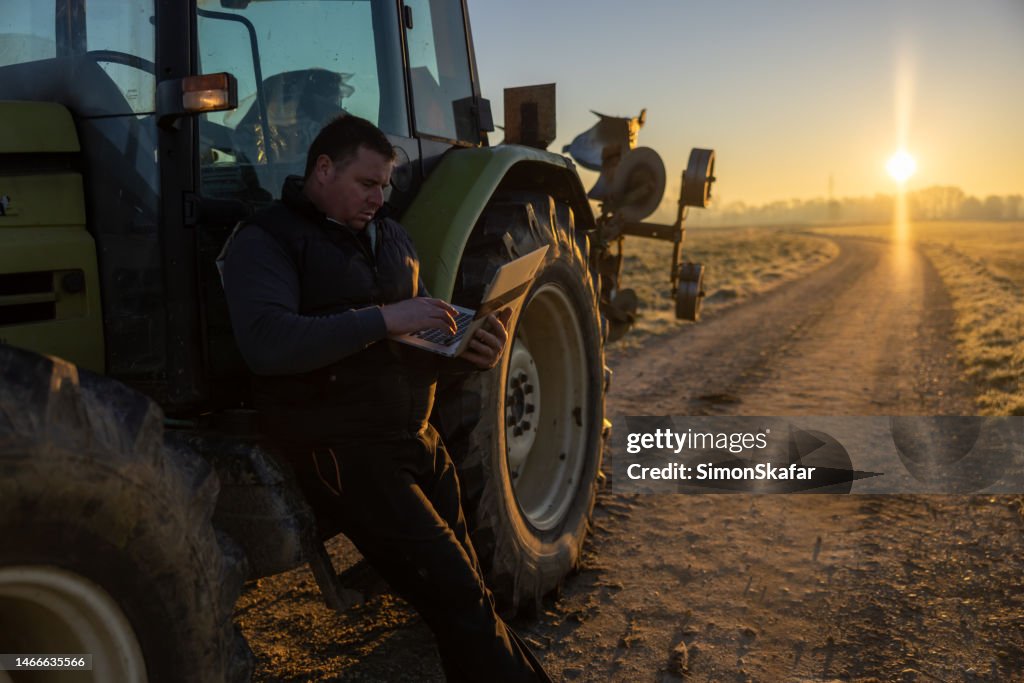 Man with short brown hair is using his laptop,leaning against his tractor in the evening parked on an agricultural path during sunset