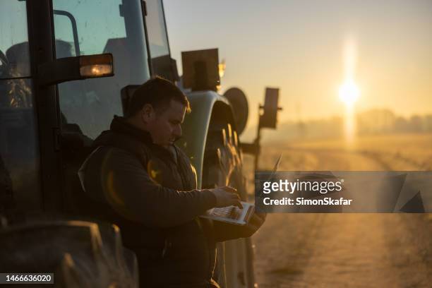male farmer with short brown hair is working on his laptop while leaning against his tractor in the evening - trucking stock pictures, royalty-free photos & images