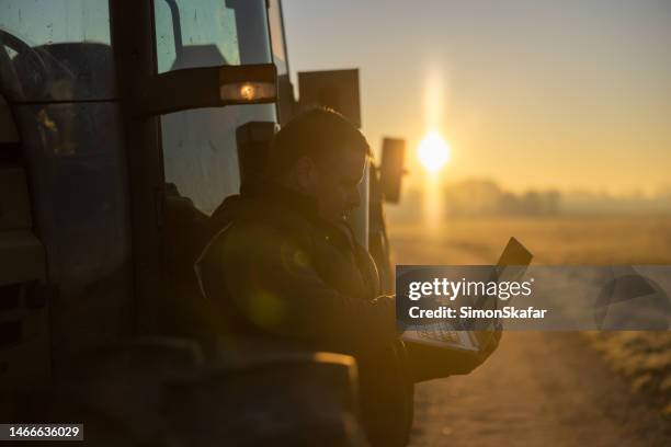 male farmer with short brown hair,planning his harvest on his laptop while leaning against his tractor,parked on an agricultural path in the evening during sunset - farmer dawn stock pictures, royalty-free photos & images