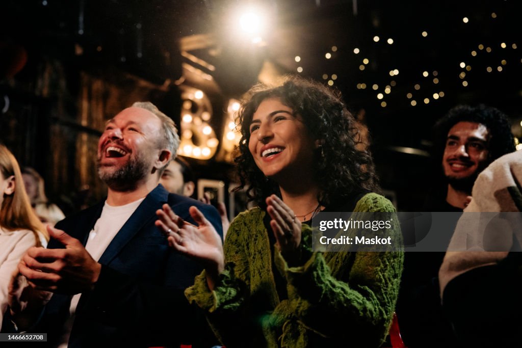 Happy audience laughing and clapping while watching comedy stage show in illuminated theater