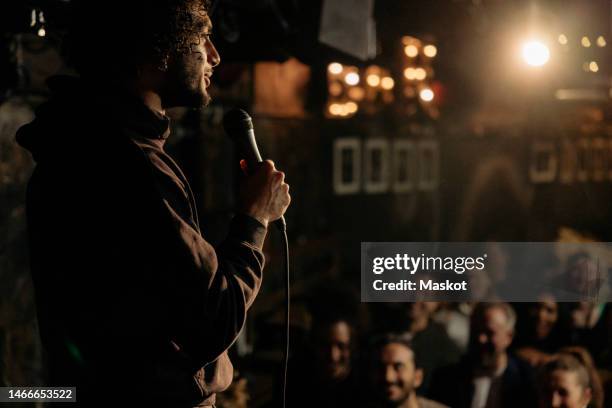 young male comedian performing stand-up for audience at illuminated amateur theater - komiek stockfoto's en -beelden