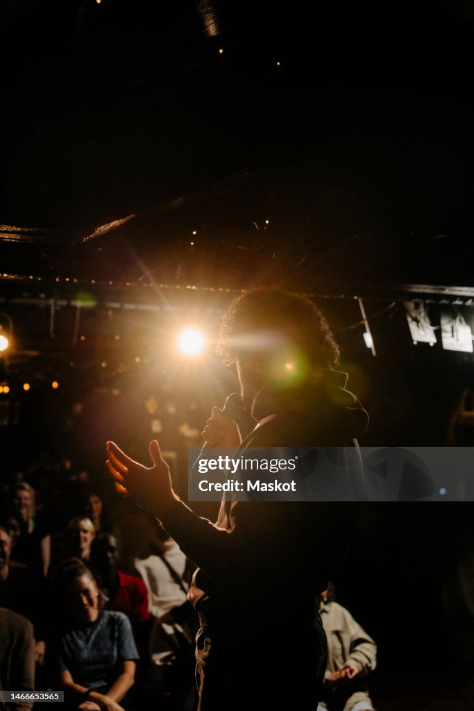 Young man performing stand-up comedy for audience at illuminated stage theater