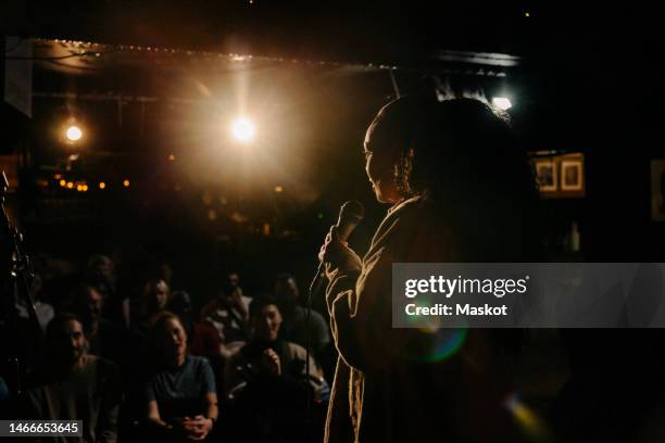 young woman performing stand-up comedy for multiracial audience at illuminated stage theater - funny people fotografías e imágenes de stock