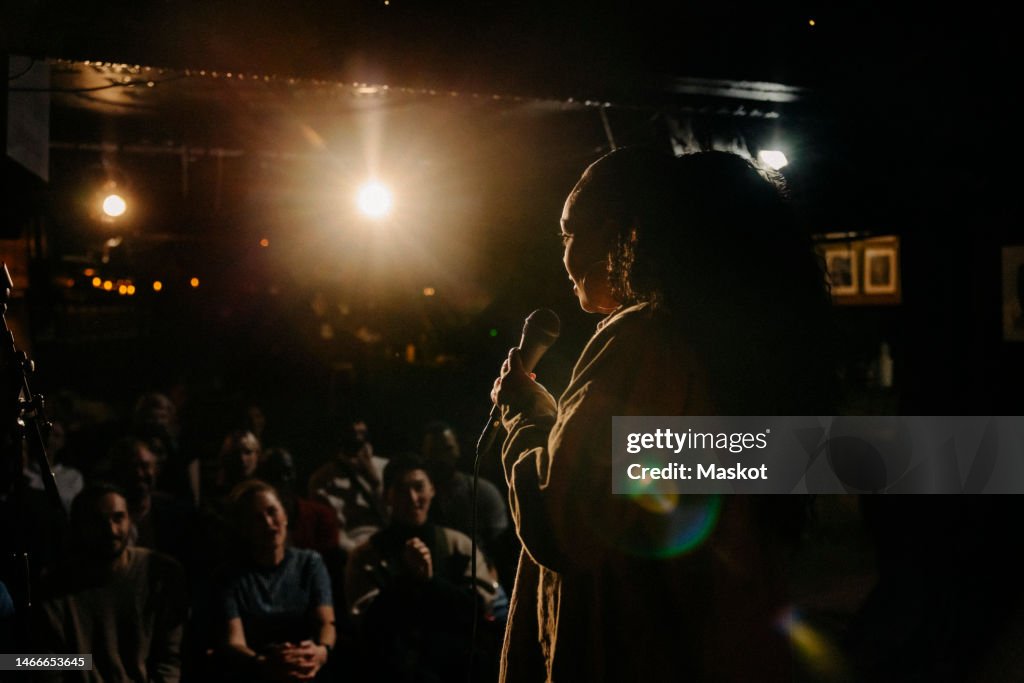 Young woman performing stand-up comedy for multiracial audience at illuminated stage theater