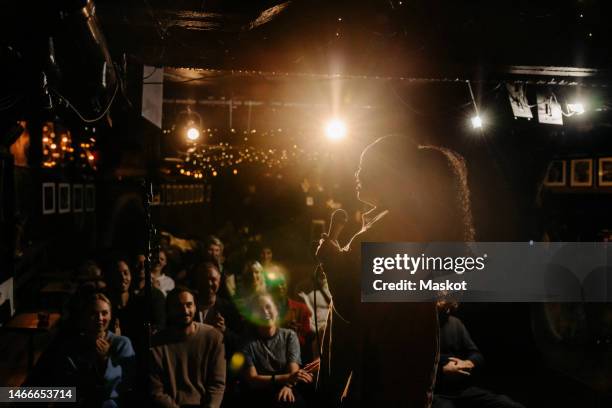 young female stand-up comedian performing for multiracial audience at illuminated amateur theater - komiek stockfoto's en -beelden