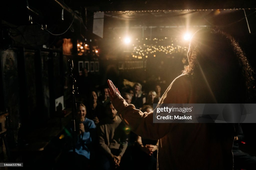 Female stand-up comedian performing for multiracial audience at illuminated stage theater