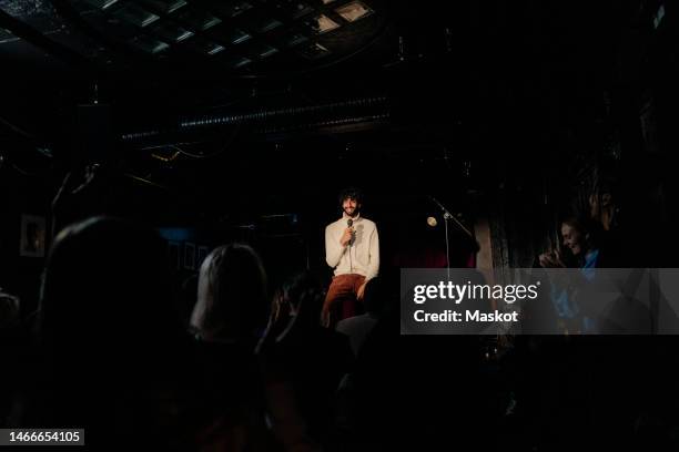 smiling young male comedian performing stand-up in front of audience on stage at amateur theater - microphone stand stock-fotos und bilder