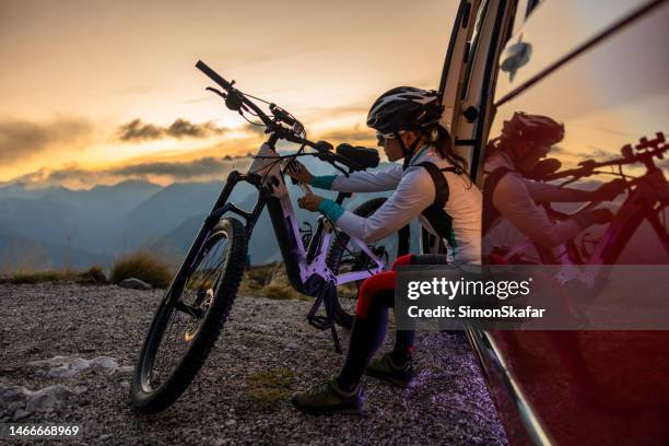 female cyclist with long brown hair wearing helmet while plugging in a cable to charge her e-bike on top of a mountain,sitting in a red van - e sport stock pictures, royalty-free photos & images