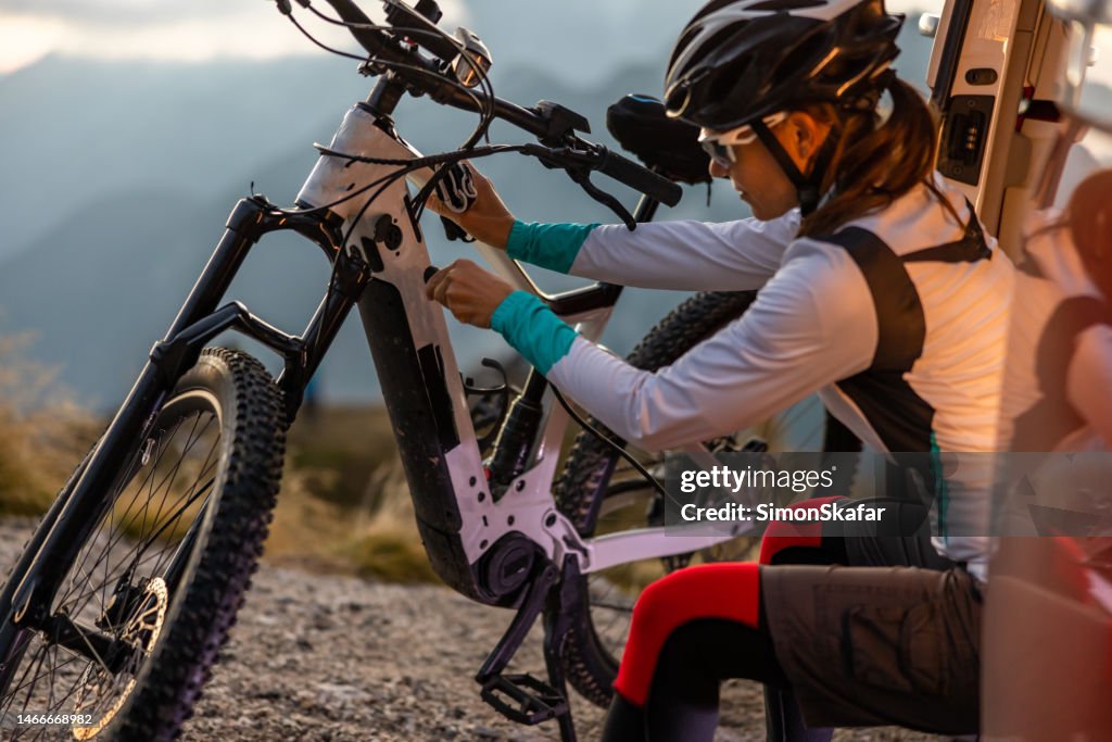 Adult woman with long brown hair wearing a helmet while plugging in a cable to charge her e-bike during a break from her biking trip to the top of Mangart