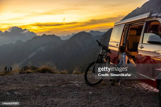 adult woman with long brown hair taking a break from her biking trip,sitting inside a van with opened door and looking at the view,bike standing in front of her during sunset - sitting on top of car stock pictures, royalty-free photos & images
