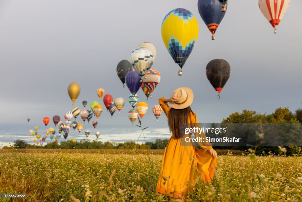 Woman with brown hair holding her hat while looking at hot air balloons flying in the air over agricultural field during sunrise