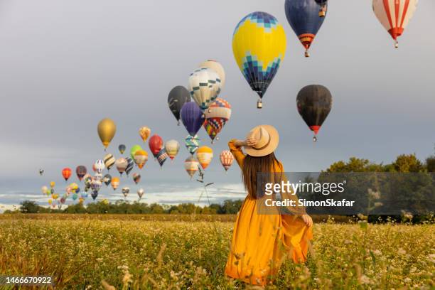 mulher com cabelos castanhos segurando seu chapéu enquanto olha para balões de ar quente voando no ar sobre o campo agrícola durante o nascer do sol - multi colored dress - fotografias e filmes do acervo