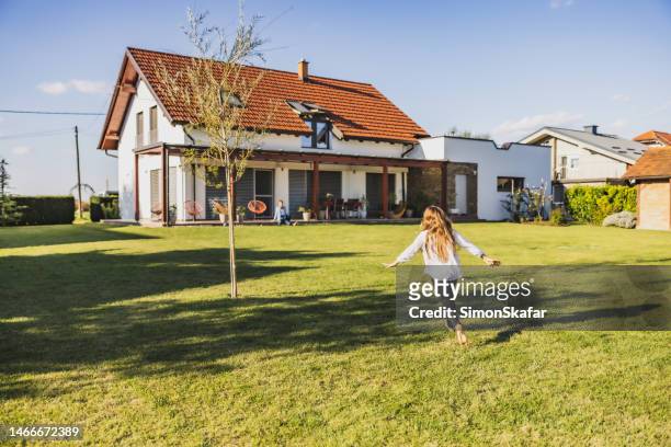 little girl is running through the garden on lawn with a tree next to her,spreading her arms while running,house in the background - small garden stock pictures, royalty-free photos & images