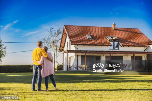 couple, homme et femme, s’étreignant tout en se tenant sur la pelouse dans la cour arrière de leur nouvelle maison achetée, vue arrière, maison moderne en arrière-plan - buy house photos et images de collection